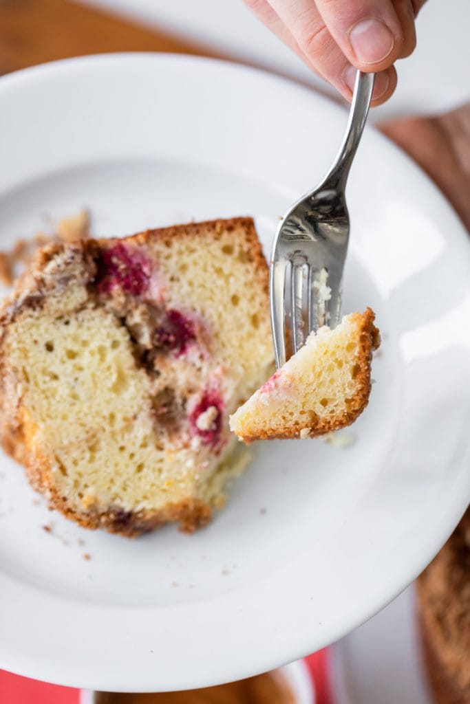 Hand holding fork with piece of coffee cake on it with rest of piece on white plate in background