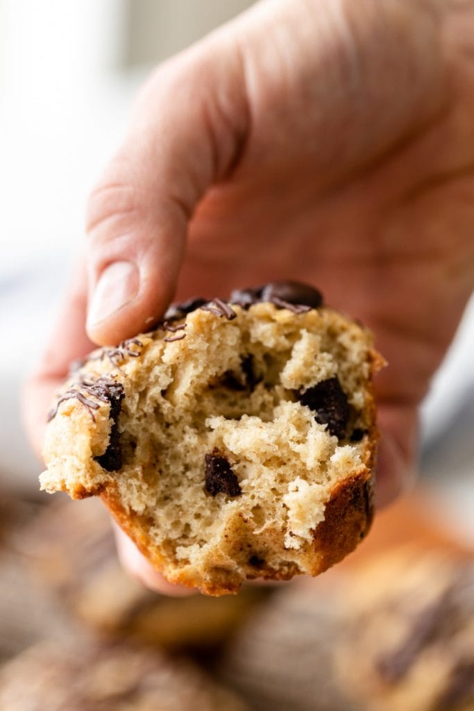 Banana muffin ripped in half being held by hand with chunks of chocolate inside with extra muffins in background