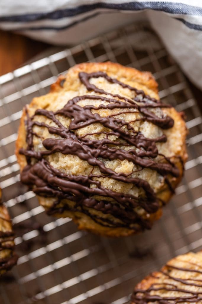 Top down view of banana muffin drizzled with chocolate sauce sitting on wire cooling rack with other muffins and towel surrounding the muffin