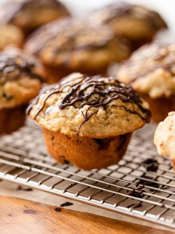 Banana chocolate chunk muffin sitting on wire cooling rack on wood board surrounded by other muffins all drizzled with chocolate on top