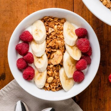 Top down view of granola in bowl on top of yogurt nestled among raspberries and bananas all on wood surface
