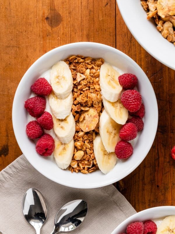 Top down view of granola in bowl on top of yogurt nestled among raspberries and bananas all on wood surface