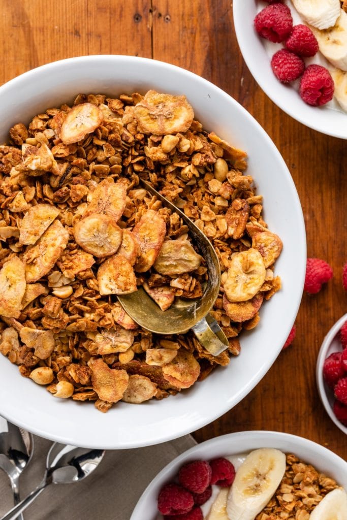 Top down view of granola sitting in white bowl surrounded by bowls of yogurt topped with granola and raspberries and bananas on wood surface
