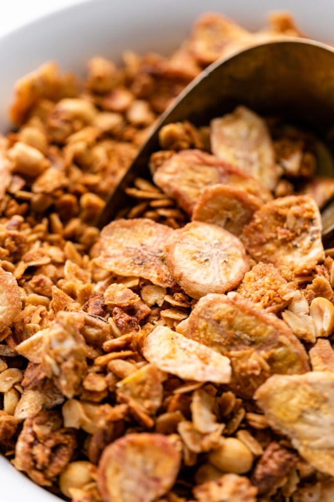 Granola with pieces of dried banana chips sitting in white bowl with gold scoop in background