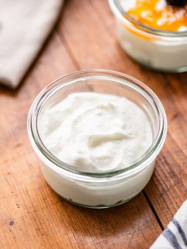 Small glass jar filled with homemade yogurt on wooden surface with second bowl in background with jam on top.