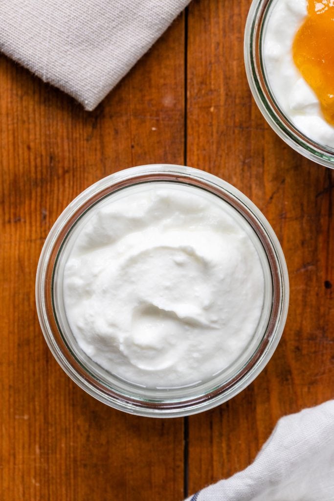 Top down view of yogurt in glass container with no toppings sitting on wood board with towel in corner