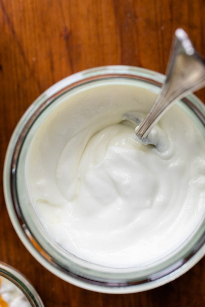 Top down view of yogurt container filled with yogurt with spoon in middle all on wood board