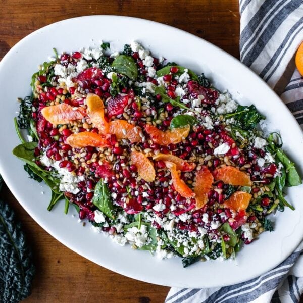 Top down view of blood orange and wheat berry salad topped with sunflower seeds and feta cheese on a white serving platter amidst kale and oranges all on a wood surface