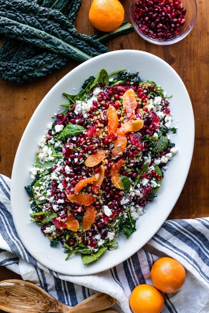 Top down view of blood orange and wheat berry salad sitting on white platter surrounded by towels and extra oranges on wood surface