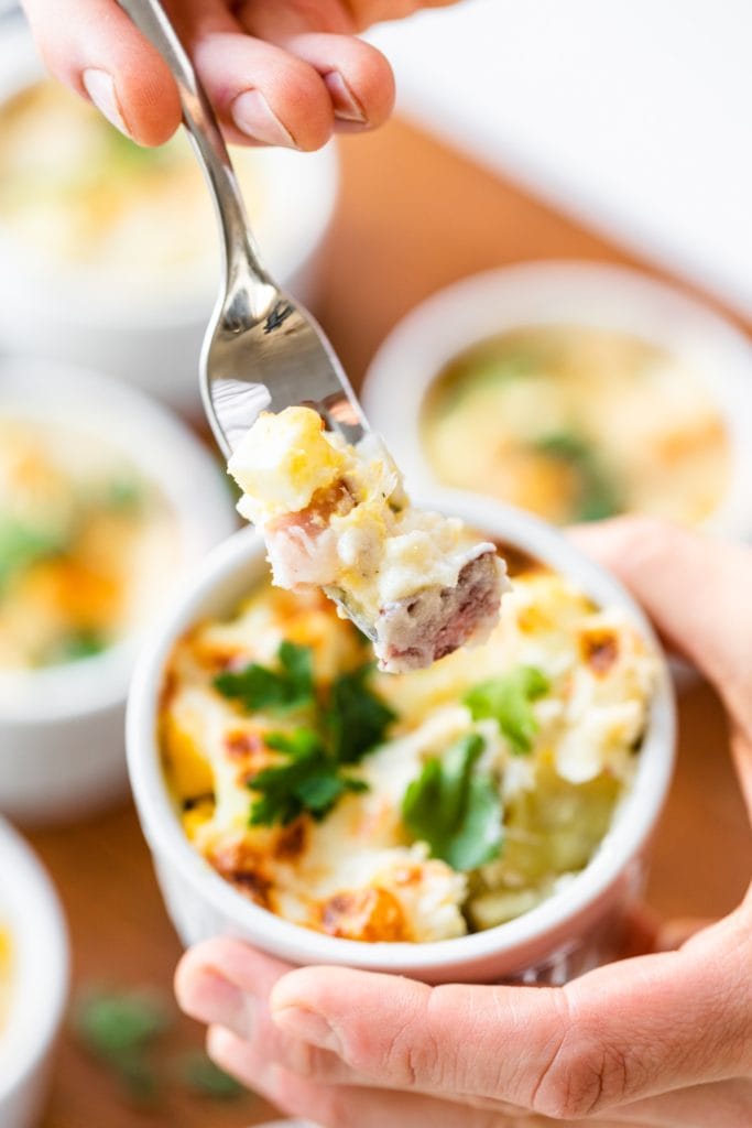 Hand holding fork full of egg, ham, and sweet potato casserole with extra casserole dishes in background