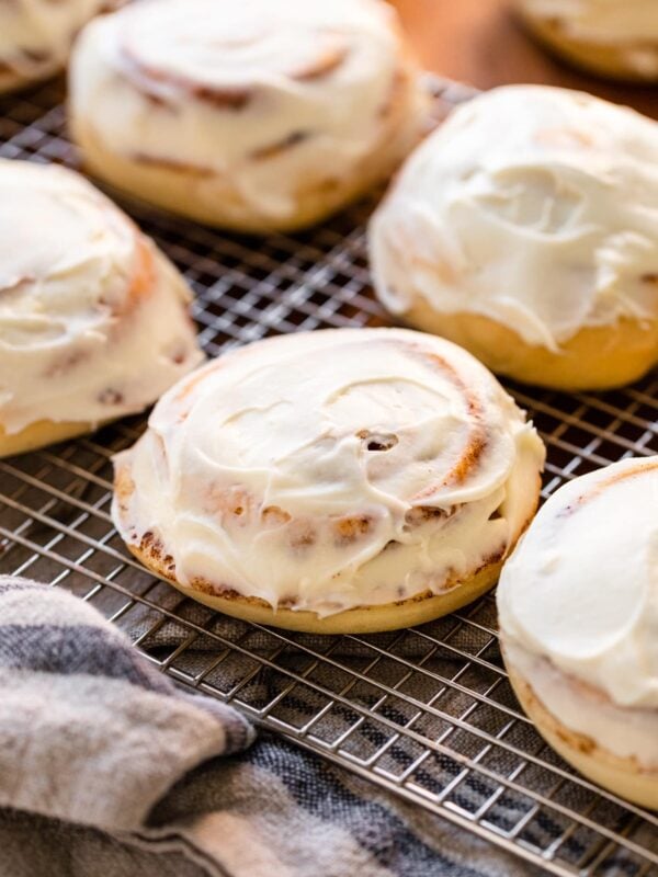 Ultimate cinnamon roll sitting on wire cooling rack surrounded by other cinnamon rolls frosted with cream cheese frosting