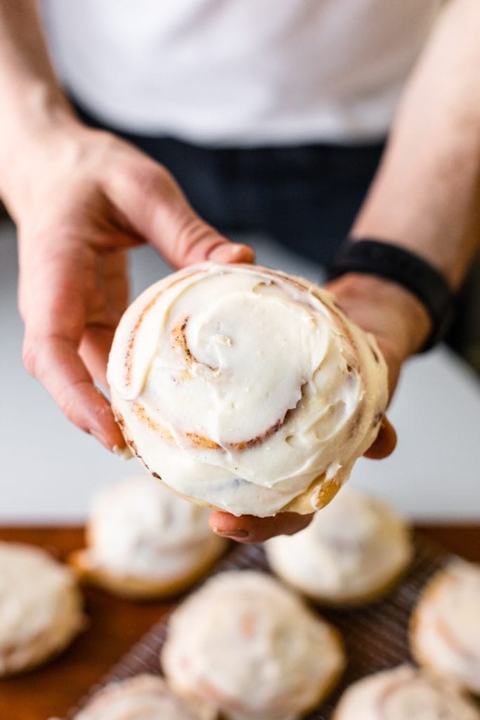 Two hands holding cinnamon roll frosted with cream cheese frosting with extra cinnamon rolls cooling in background