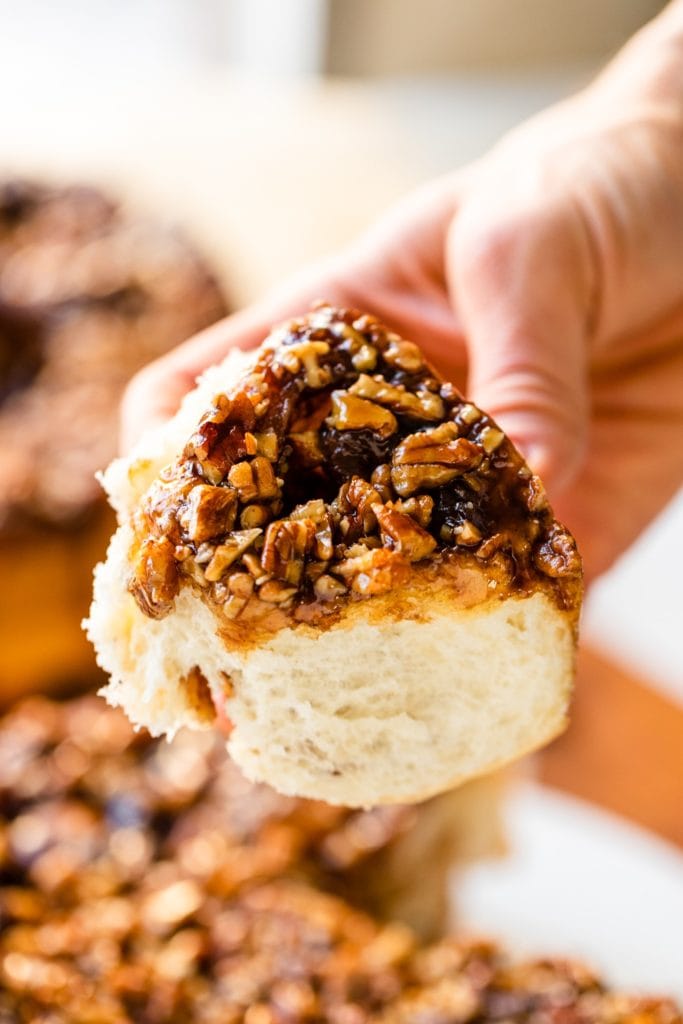 Hand holding cinnamon roll topped with sticky caramel sauce with chopped pecans and caramel sauce on top with extra rolls in background