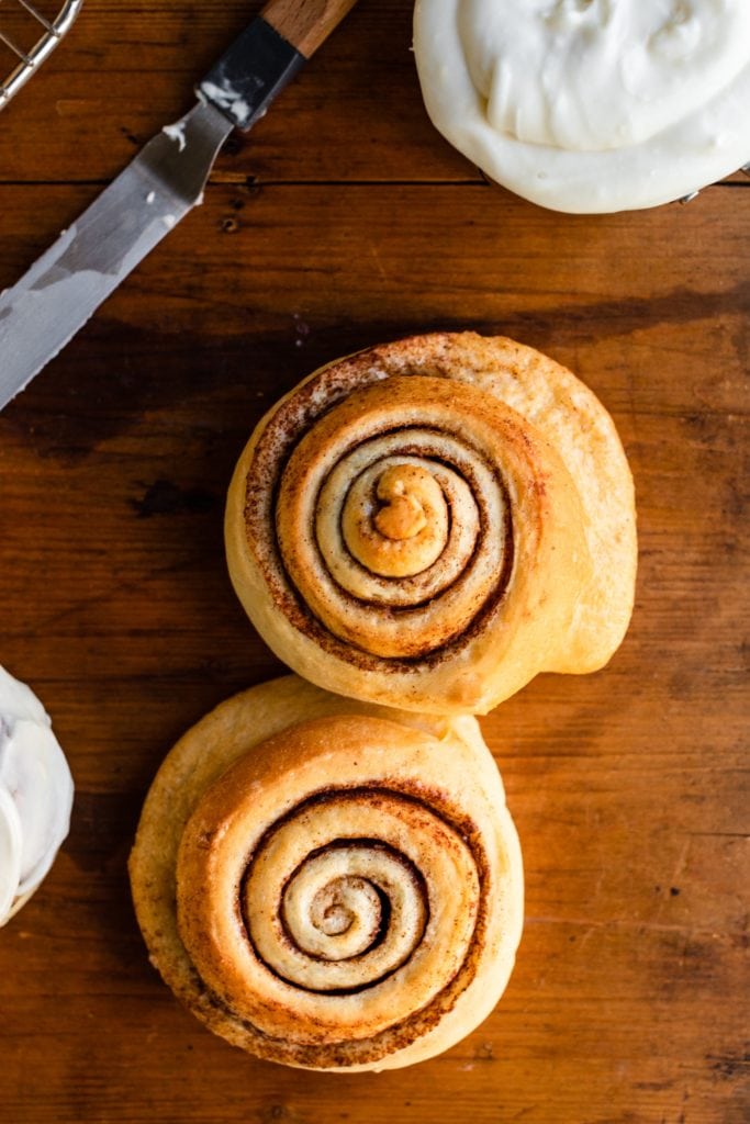 Top down view of unfrosted cinnamon rolls sitting on wood surface with frosting in measuring cup and knife ready to frost