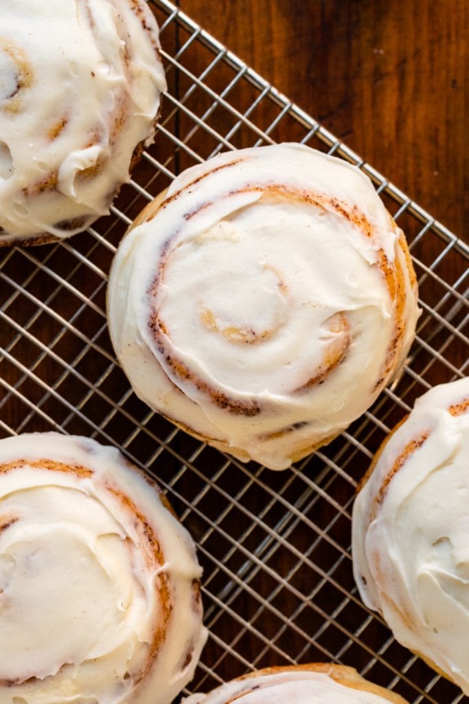 Top down view of ultimate cinnamon roll recipe sitting on wire cooling rack surrounded by other cinnamon rolls all frosted with white cream cheese frosting all on wood surface