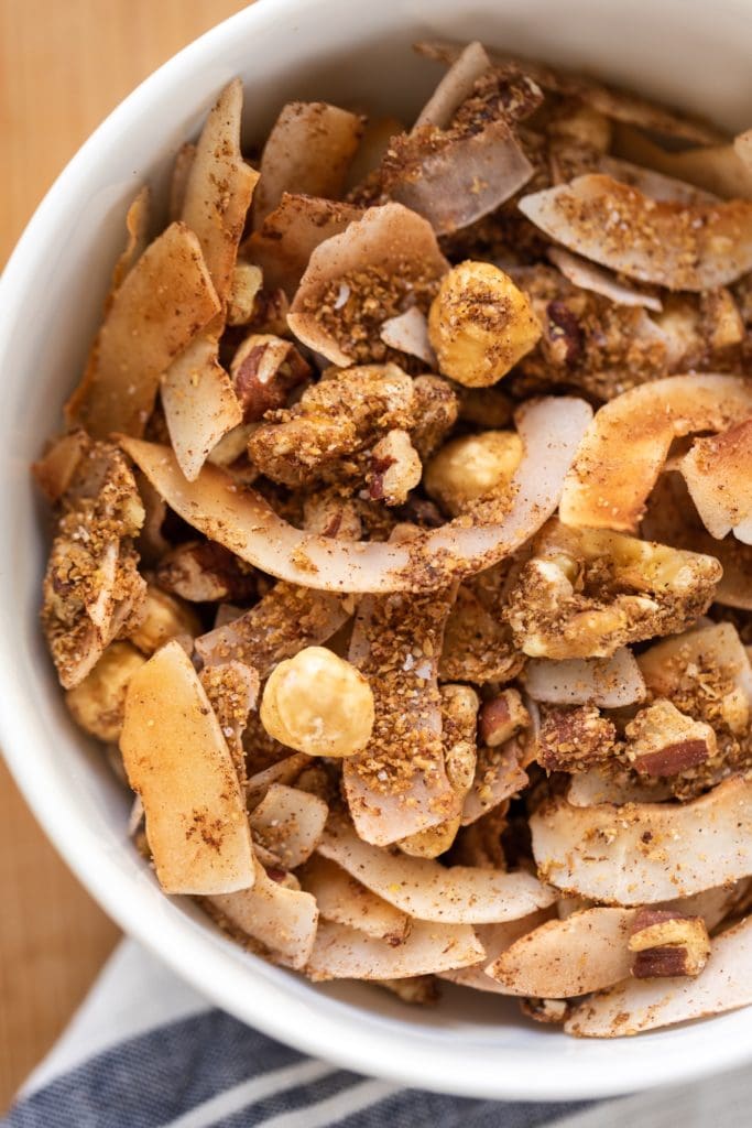 Coconut and nut granola sitting in white bowl with white and blue linen napkin all on wood surface