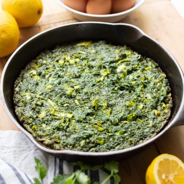 Herb omelet stuffed with greens sitting in skillet with lemons and eggs beside on wood surface