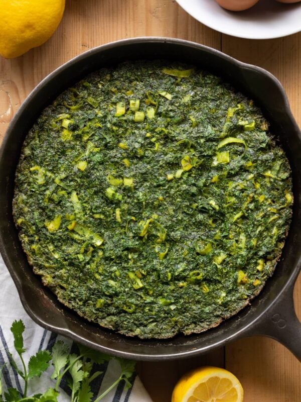 Top down view of overstuffed herb omelet in a skillet with lemons and eggs in white bowl all on wood surface