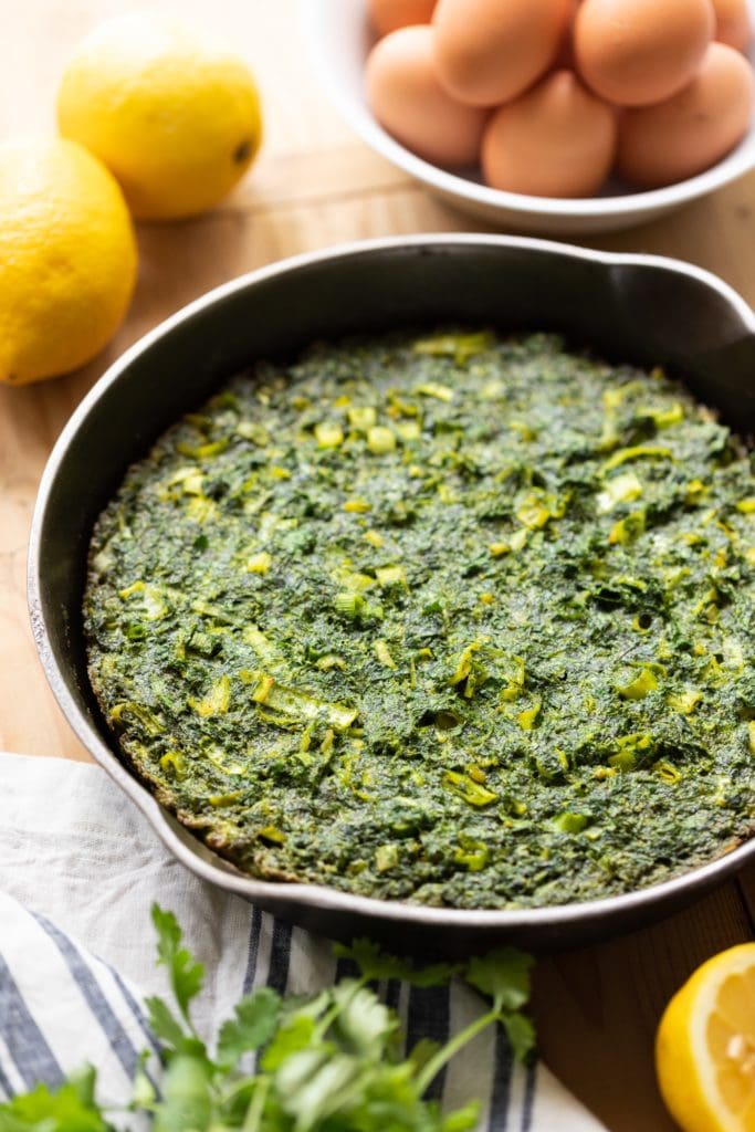Cast iron skillet filled with omelet dotted with leeks with lemons and eggs in background all on wood surface