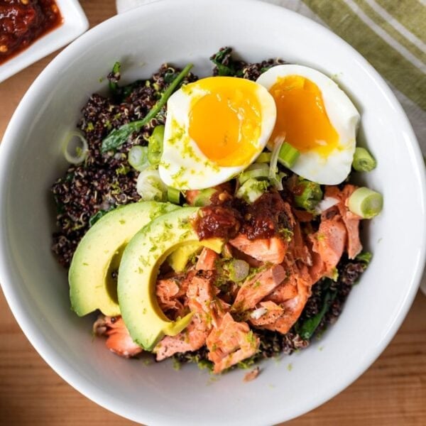 Top down view of salmon bowl topped with avocado and jammy eggs all on wood surface with toppings set around