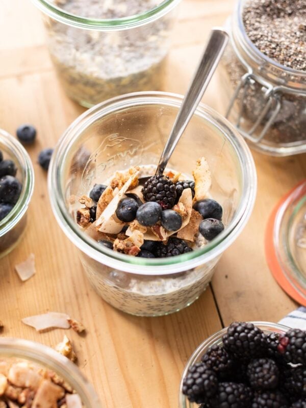 Vegan overnight oats sitting in glass container topped with granola and blueberries all on wood surface with extra toppings spread around