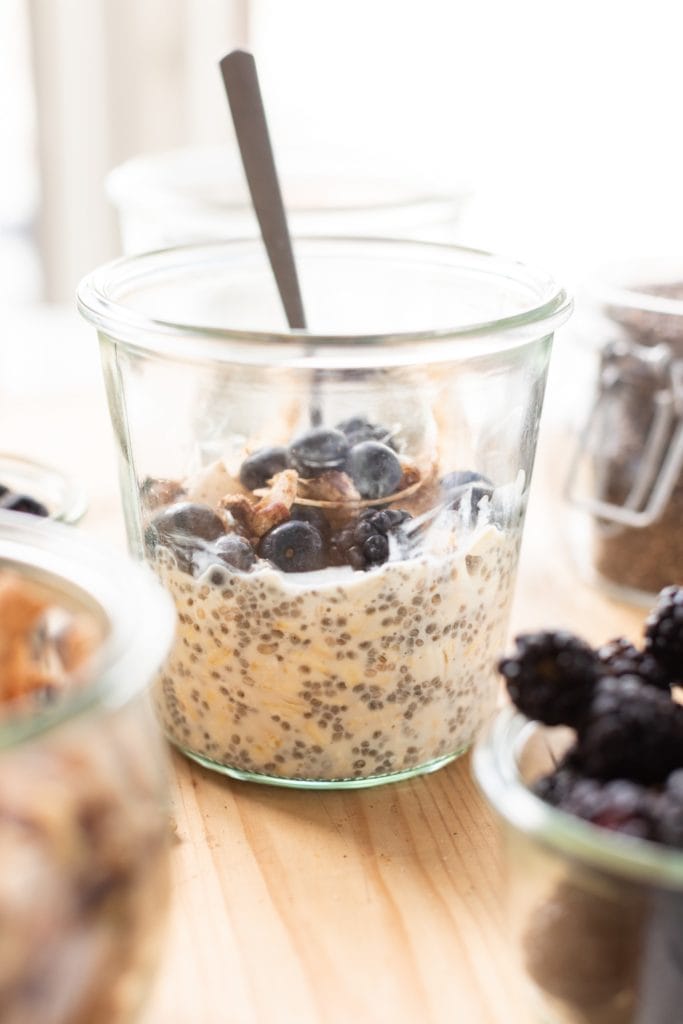 Glass container filled with overnight oats and topped with blueberries and granola with a spoon all on wood surface