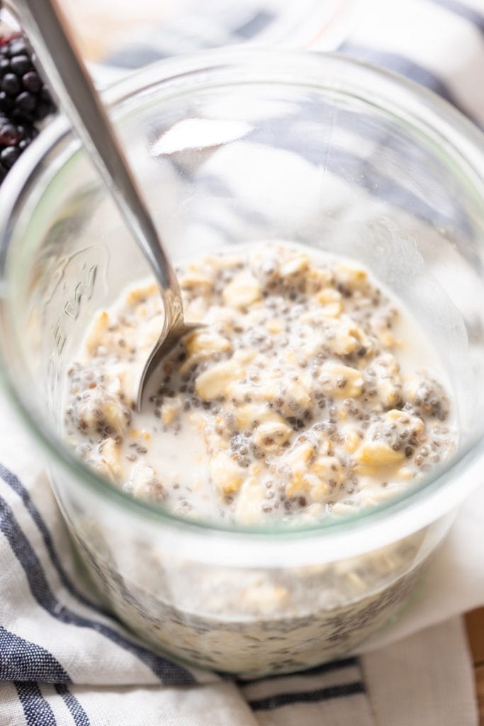 Vegan overnight oats sitting in glass container with spoon resting on side of container all on a striped linen towel