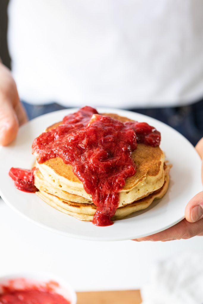 Two hands holding plate full of pancakes smothered with bright red strawberry rhubarb sauce with white shirt in background