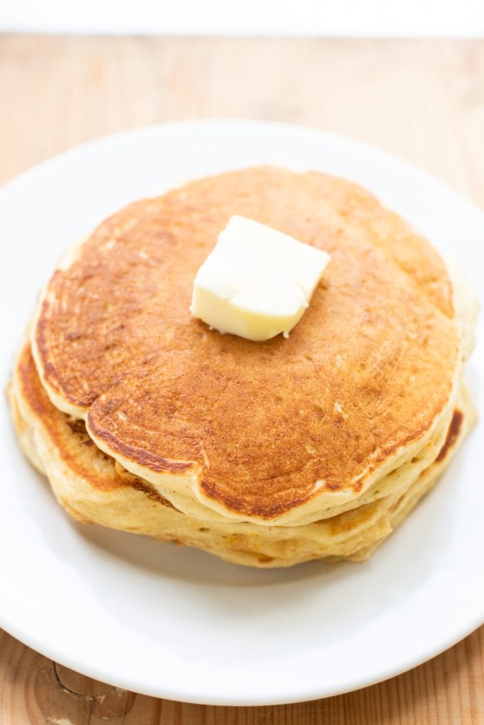 Three pancakes stacked on top of each other with pat of butter on top all on white plate sitting on wood surface