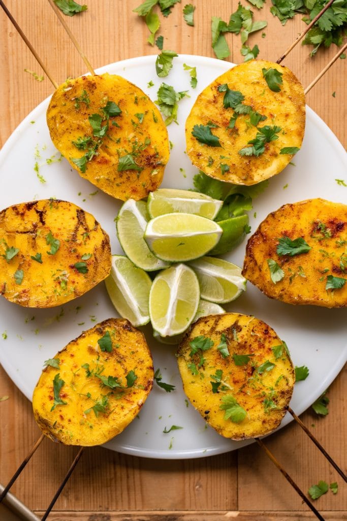 Top down view of six grilled chili mangoes sitting on circular white serving dish topped with cilantro and spices with limes in the middle all on wood surface
