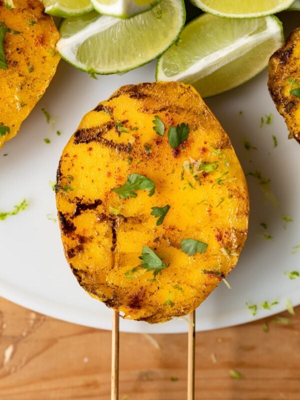 Top down view of grilled mango sitting on white serving dish on wood table sprinkled with cilantro and spices surrounded by limes and other mango