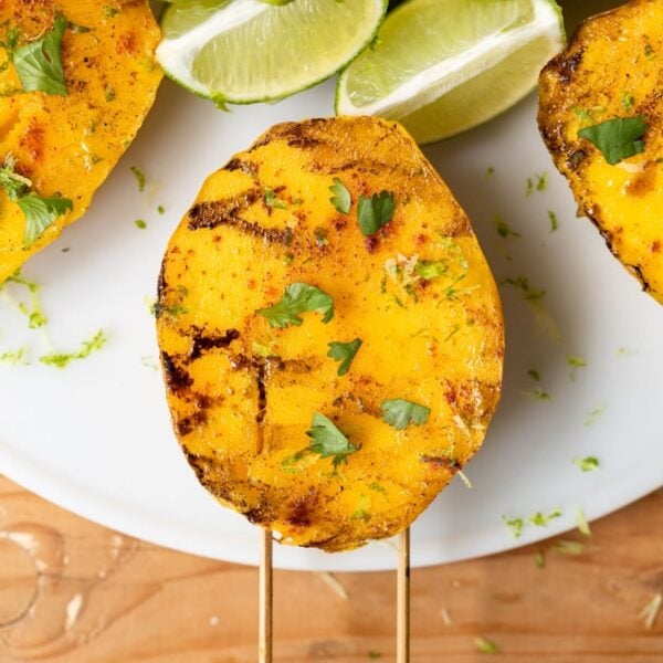 Top down view of three mangoes with grill marks and sprinkled with spices and cilantro sitting on white plate on a wood table with limes