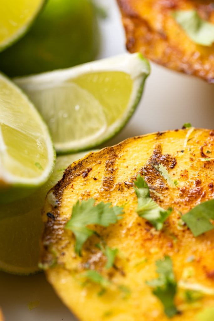 Close up view of mango sitting on white surface with limes and topped with cilantro and spices with grill marks