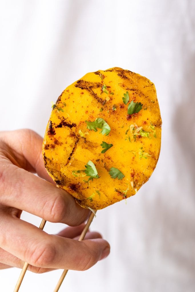 Hand holding grilled chili mango topped with cilantro and spices with two sticks coming out of bottom in front of white t-shirt