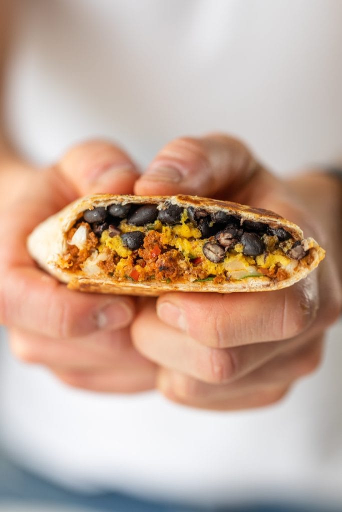 Two hands holding southwestern breakfast burrito with layers of black beans, chorizo, cheese, and eggs with white shirt in background