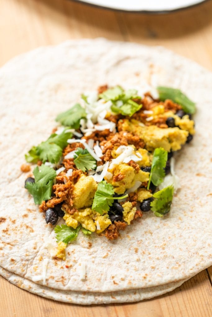 Open tortilla with layers of chorizo, eggs, and black beans sprinkled with cilantro, cheese, and onions on wood surface