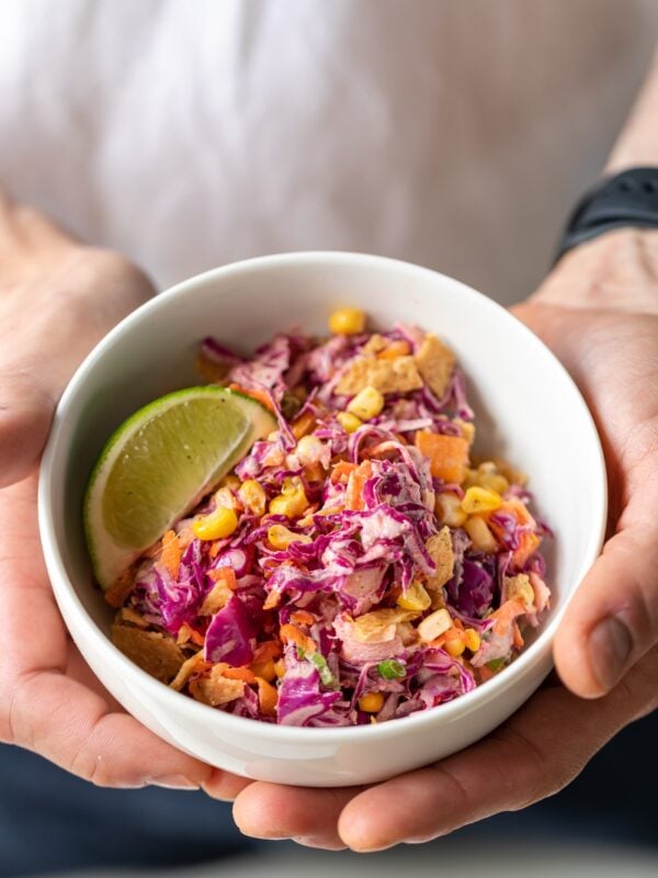 Two hands holding white bowl filled with Tex-Mex coleslaw made with red cabbage served with a lime wedge and topped with tortilla chips