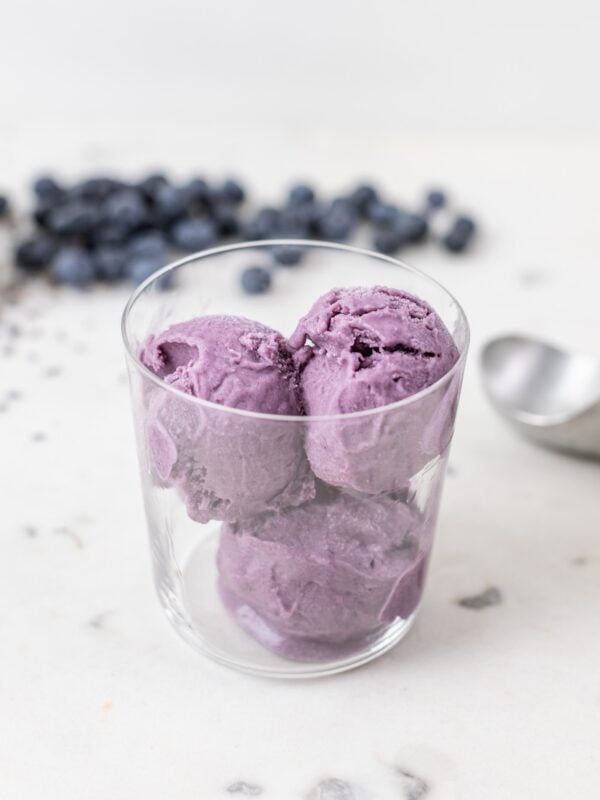 Three scoops of blueberry lavender ice cream sitting in glass container with blueberries and ice cream scoop in background all on marble surface
