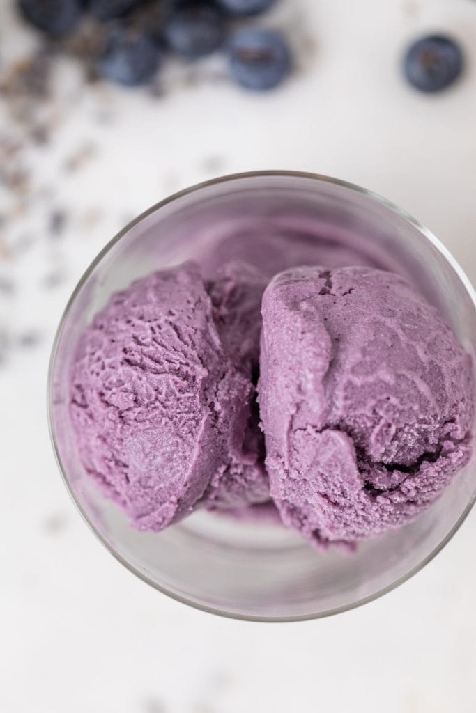 Top down view of purple ice cream in glass container with blueberries and dried lavender on marble surface