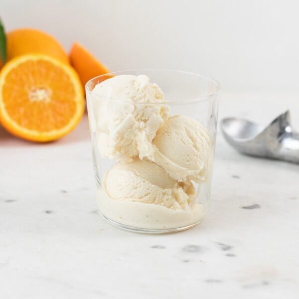 Three scoops of brown butter orange sage ice cream in a glass cup with orange slices and ice cream scoop sitting on piece of marble