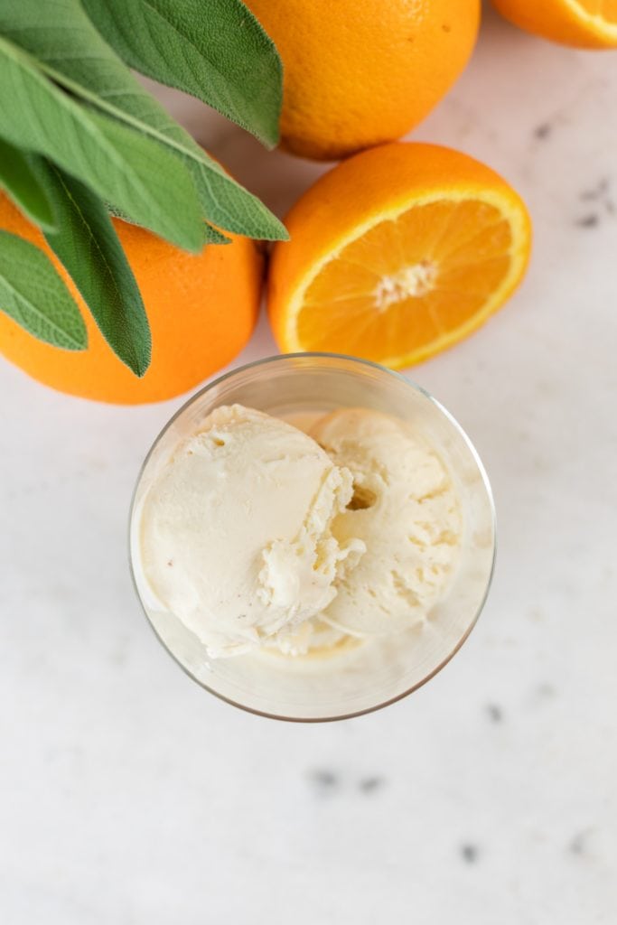 Top down view of orange sage ice cream sitting in glass container with sage leaves and orange slices all on marble surface