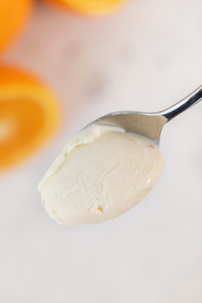 Spoon holding spoonful of white ice cream with orange slices in background with marble surface in background