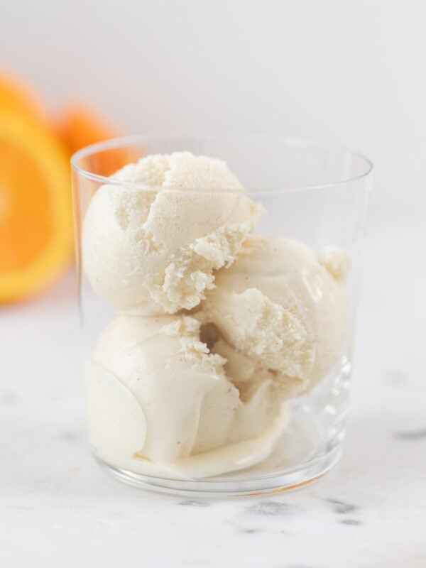 Three scoops of brown butter orange sage ice cream sitting in glass cup with sliced oranges in background all on marble surface