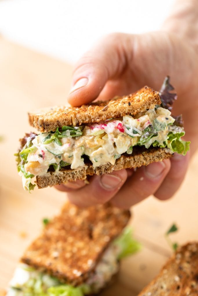 Hand holding a sandwich filled with chickpea salad and topped with lettuce and radish with extra half of sandwich sitting on wood board in background