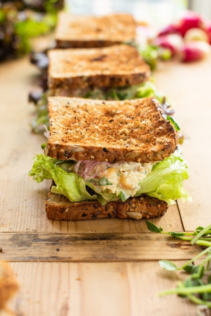 Chickpea salad sandwiched in between two toasted pieces of bread with lettuce and radishes sitting on wood board with extra sandwiches in background