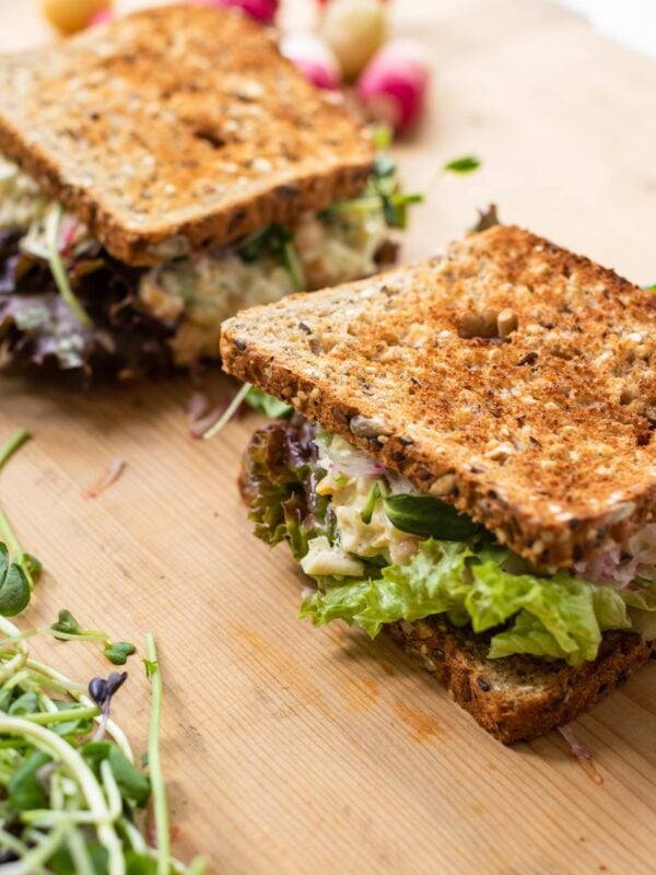 Wood cutting board with two toasted sandwiches filled with lettuce and chickpea salad with radish in background