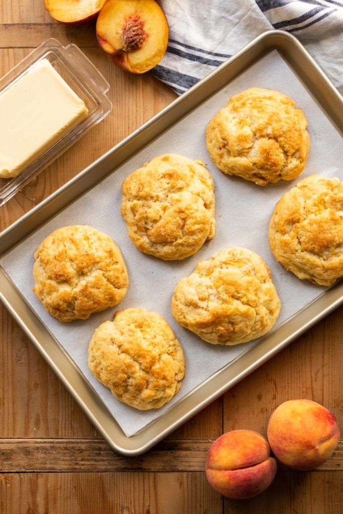 Top down view of peach drop biscuits cooling on piece of parchment paper in a baking tin with butter and sliced peaches in background all on wooden surface
