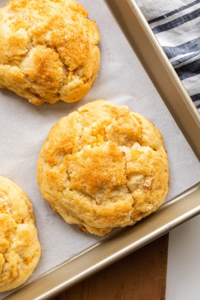 Three golden brown biscuits on piece of white parchment sitting in a baking dish with striped linen in background all on wooden surface