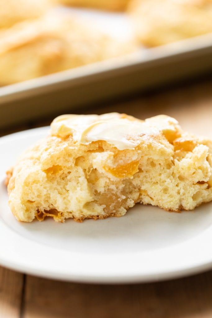 Peach drop biscuit cut in half with center exposed slathered with butter on white plate with extra biscuits on baking sheet in background all on wooden surface