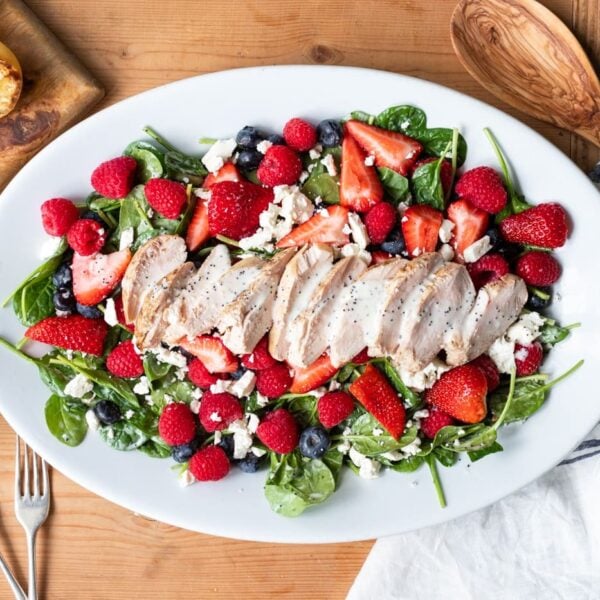 Top down view of grilled chicken berry salad sitting on bed of spinach on white plate surrounded with squeezed lemons, wooden spoons, and linen all on wood surface